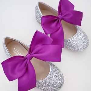 May include: A pair of silver glitter ballet flats with a large purple satin bow on each shoe. The shoes have a simple design and are perfect for a special occasion.