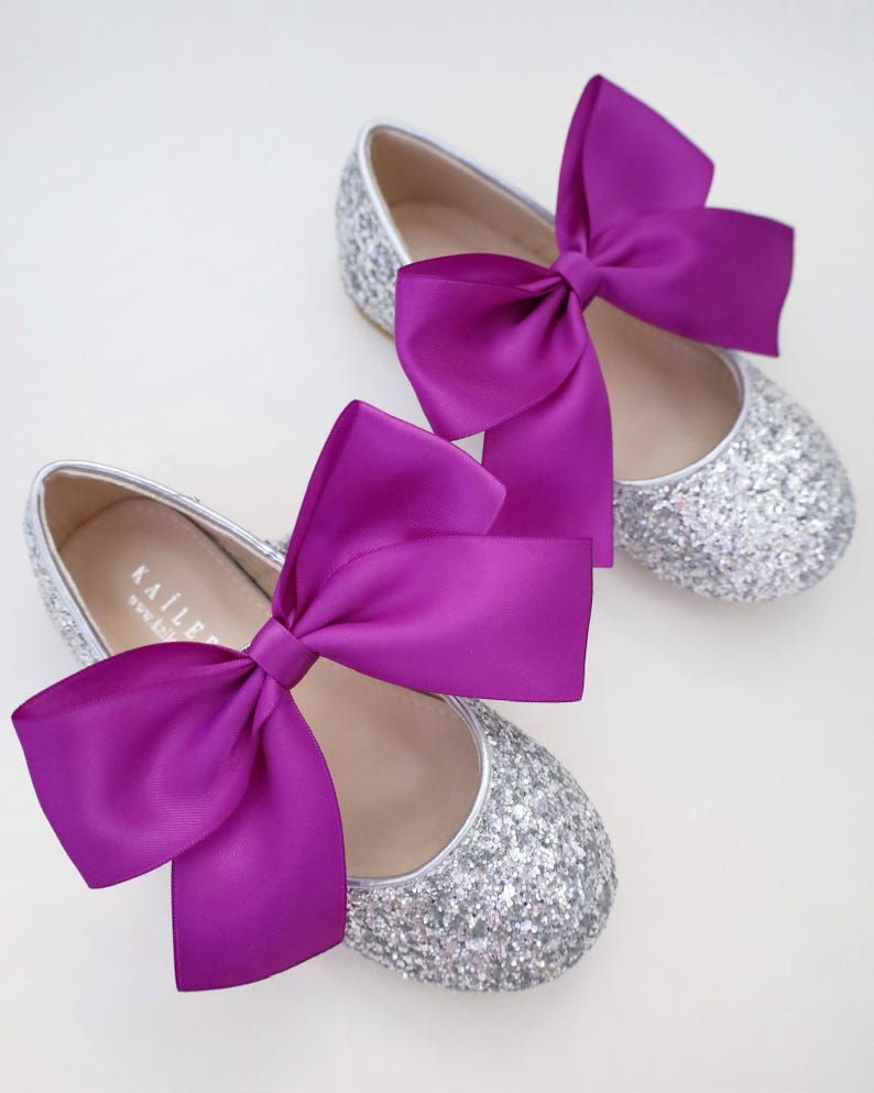 May include: A pair of silver glitter ballet flats with a large purple satin bow on each shoe. The shoes have a simple design and are perfect for a special occasion.