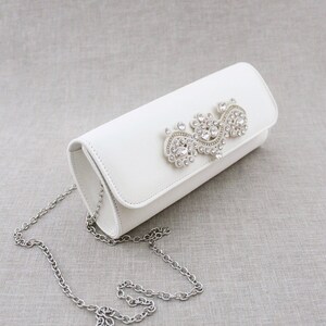 May include: White satin clutch bag with a silver chain strap and a large rhinestone embellishment.
