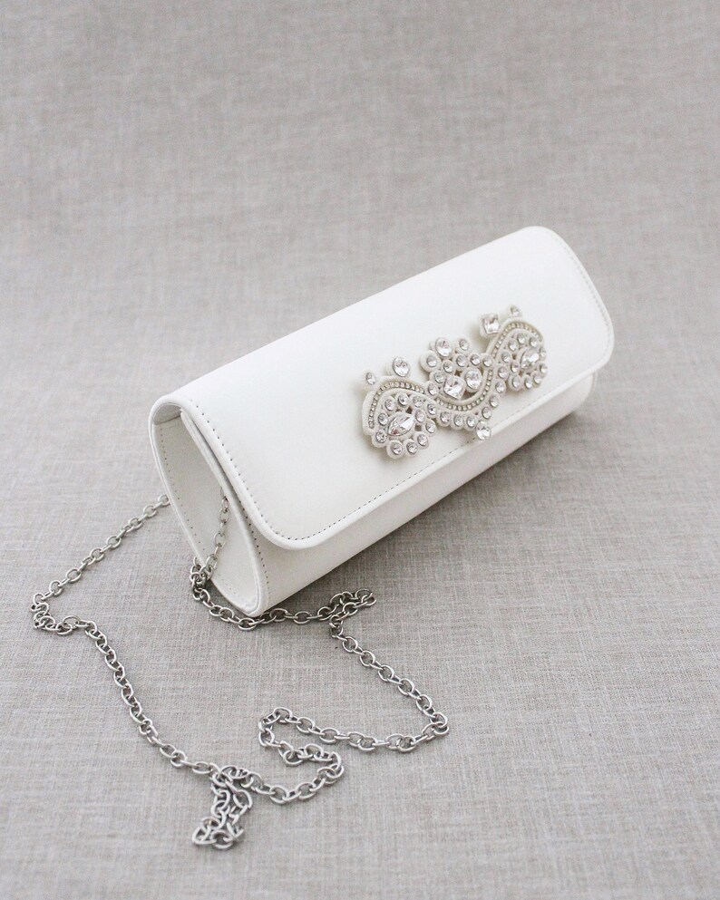 May include: White satin clutch bag with a silver chain strap and a large rhinestone embellishment.