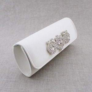 May include: A white clutch purse with a crystal embellishment in the shape of a swirl.