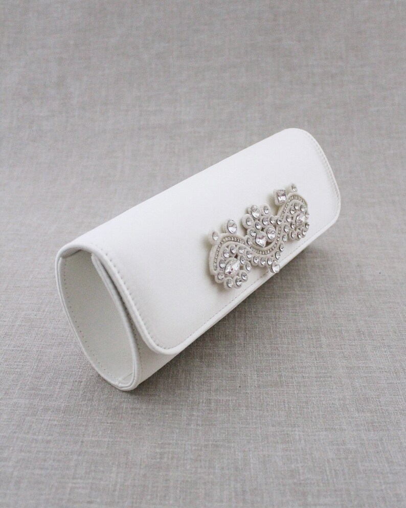 May include: A white clutch purse with a crystal embellishment in the shape of a swirl.