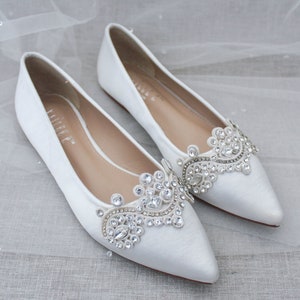 May include: Pair of white satin bridal flats with pointed toes and a jeweled embellishment on the vamp. The embellishment features a delicate floral design with clear rhinestones.