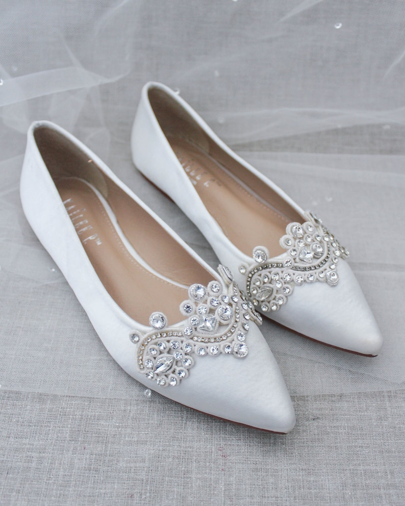 May include: Pair of white satin bridal flats with pointed toes and a jeweled embellishment on the vamp. The embellishment features a delicate floral design with clear rhinestones.