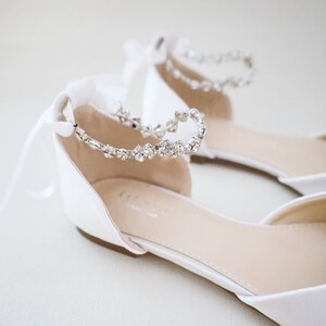 May include: A pair of white satin flats with a delicate crystal ankle strap. The shoes have a pointed toe and a low heel. White satin ribbons are tied in bows at the back of the ankle. These elegant flats are suitable for weddings or special occasions.