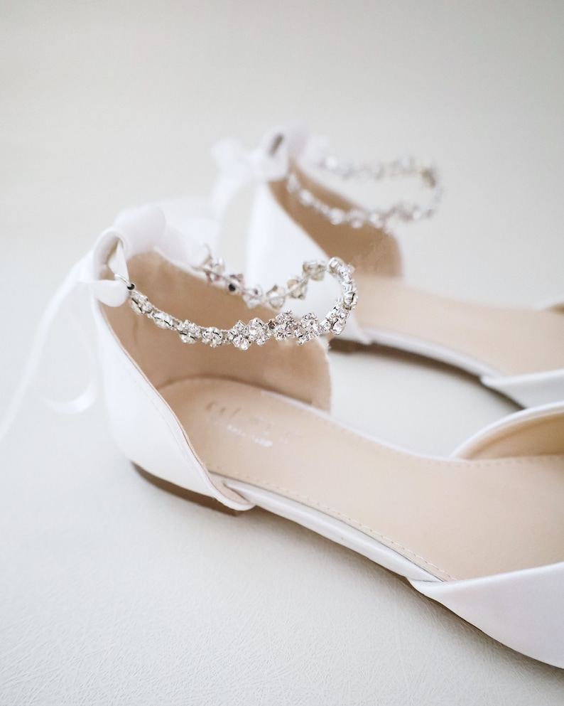 May include: A pair of white satin flats with a delicate crystal ankle strap. The shoes have a pointed toe and a low heel. White satin ribbons are tied in bows at the back of the ankle. These elegant flats are suitable for weddings or special occasions.