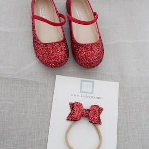Puede incluir: Zapatos rojos de purpurina con una diadema de lazo rojo. Los zapatos tienen una correa en la parte superior y la diadema es de el&aacute;stico. Los zapatos y la diadema est&aacute;n decorados con purpurina roja. La diadema tiene el texto "KP" y "www.kaileep.com" impreso en ella.