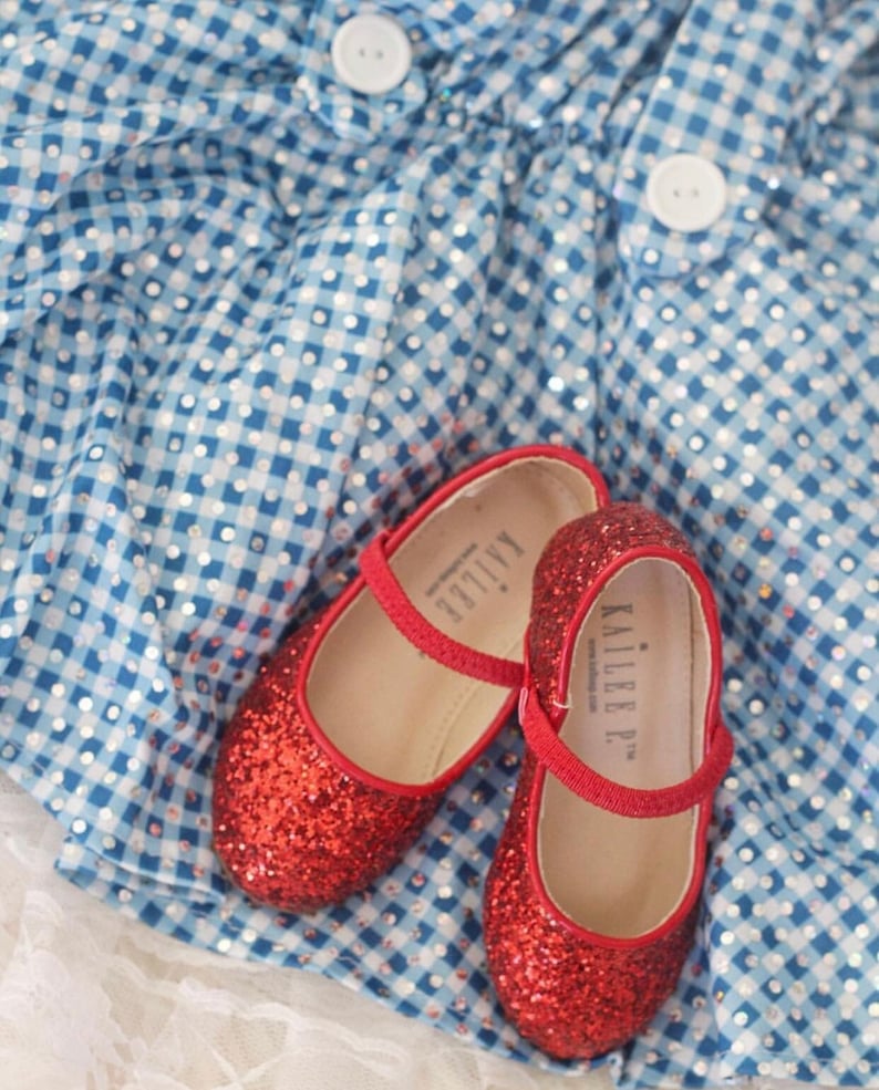 Puede incluir: Un par de zapatos rojos de purpurina con una correa y el nombre "KAILLEE P." escrito en la correa. Los zapatos est&aacute;n sobre una tela de cuadros azules y blancos.
