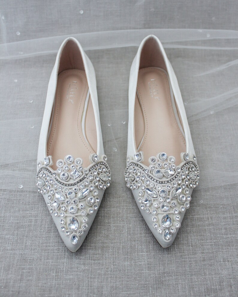 May include: A pair of white satin bridal flats with pointed toes. The shoes are embellished with a large, ornate design of clear rhinestones.