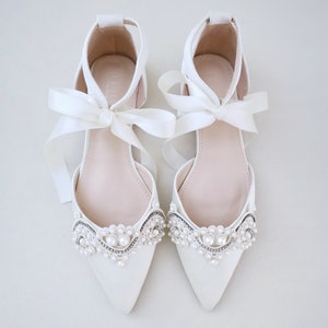 May include: A pair of ivory-colored, pointed-toe flats with delicate pearl and rhinestone embellishments. The shoes feature ankle straps and are tied with satin ribbon bows. The flats are designed for special occasions.