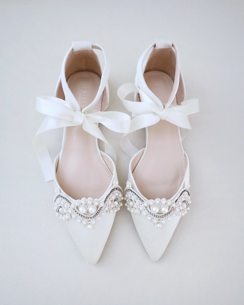 May include: A pair of ivory-colored, pointed-toe flats with delicate pearl and rhinestone embellishments. The shoes feature ankle straps and are tied with satin ribbon bows. The flats are designed for special occasions.