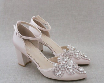 May include: A pair of blush pink satin heeled shoes with ankle straps and a pointed toe. The shoes are embellished with a large crystal design on the toe.