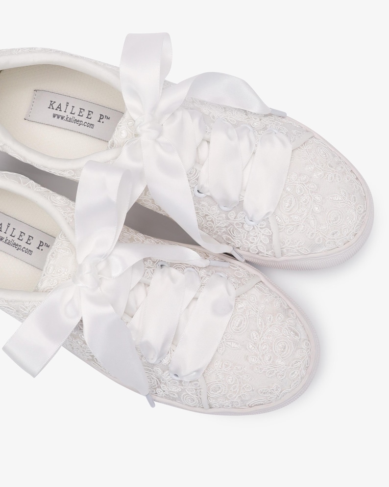 May include: White lace sneakers with satin ribbon laces. The sneakers feature a delicate floral lace pattern and a white rubber sole. The brand name "KAILEE P." is visible on the inner sole.