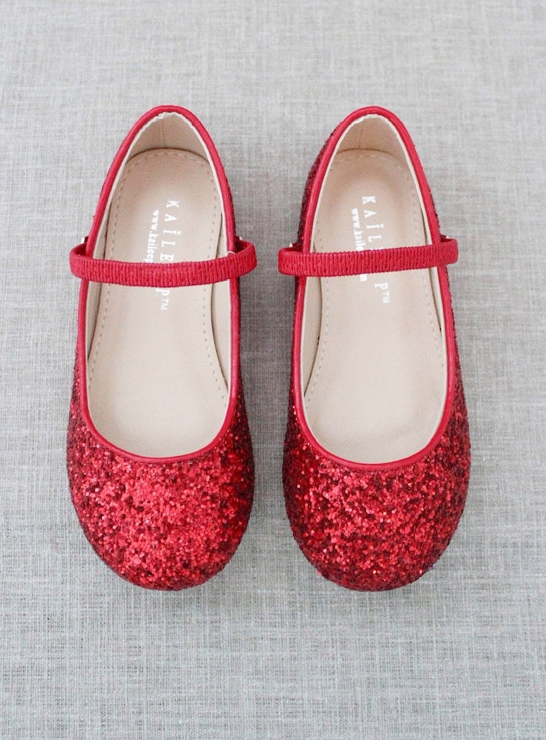 Puede incluir: Bailarinas rojas de purpurina con una tira de sat&eacute;n roja y cierre de hebilla. Los zapatos tienen una plantilla blanca con el nombre de la marca "KAILL" impreso en ella.