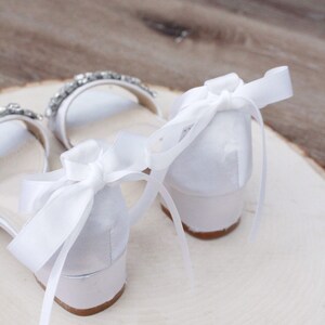 May include: White satin heeled sandals with a rhinestone embellishment and white satin ribbon ties.