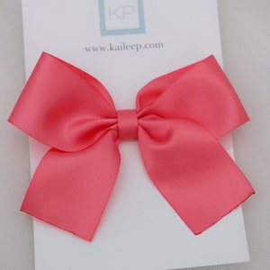 May include: A coral satin ribbon bow hair clip. The bow is tied in a classic shape with two loops and a centre knot. The hair clip is attached to the back of the bow.