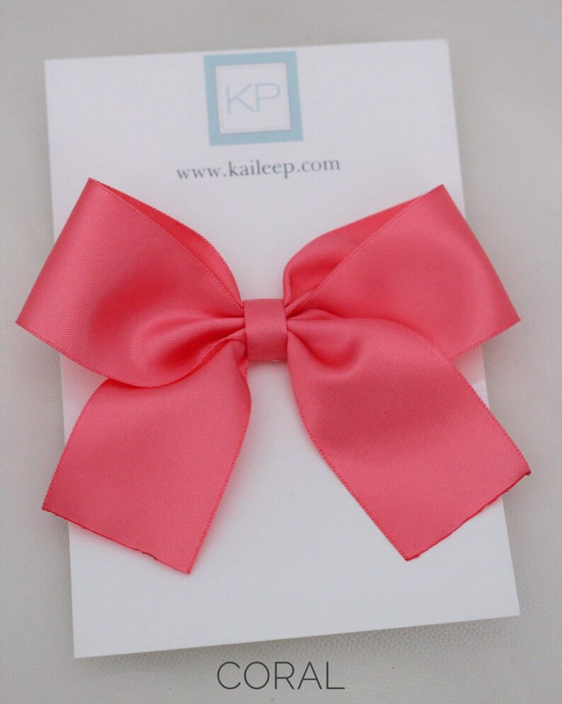 May include: A coral satin ribbon bow hair clip. The bow is tied in a classic shape with two loops and a centre knot. The hair clip is attached to the back of the bow.