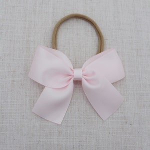 May include: A dusty pink satin bow tied with a brown elastic headband. The bow is on a light brown fabric background. Dusty Pink.