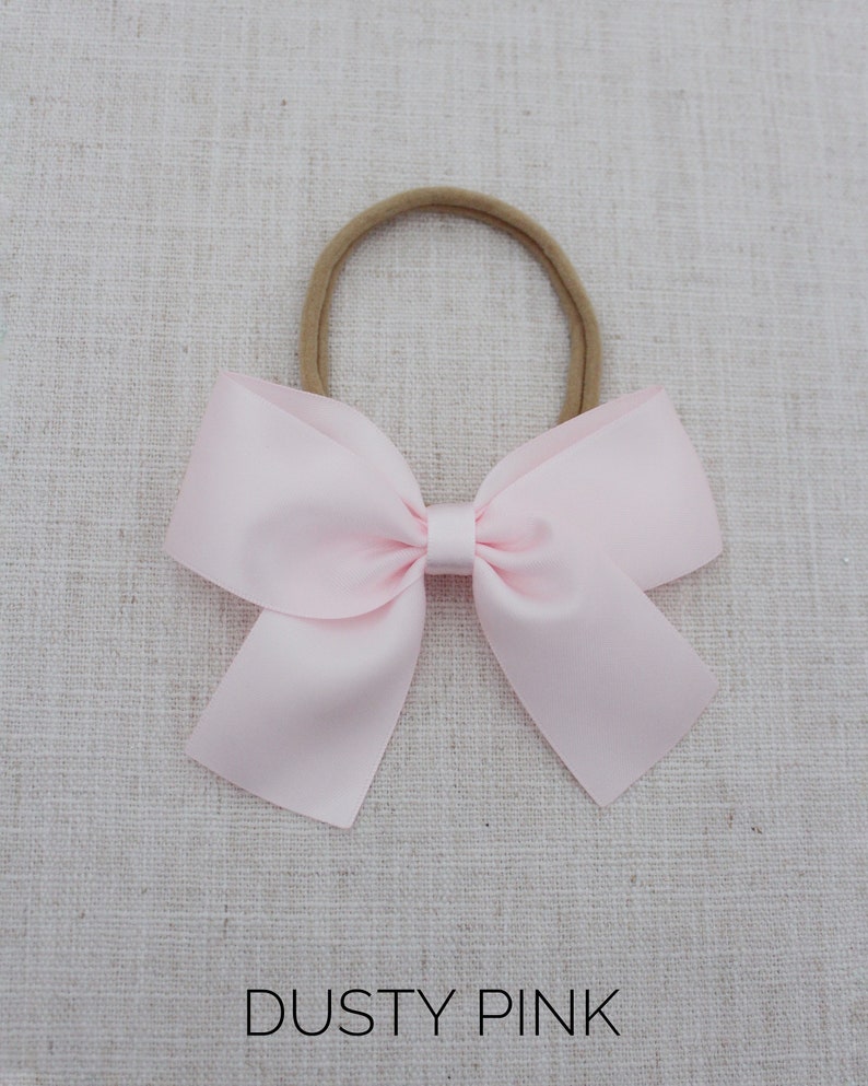 May include: A dusty pink satin bow tied with a brown elastic headband. The bow is on a light brown fabric background. Dusty Pink.