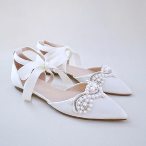 May include: A pair of ivory satin flat shoes with pointed toes. The shoes feature a decorative arrangement of pearls and rhinestones on the vamps and ankle straps tied with satin bows. The shoes are designed for special occasions.