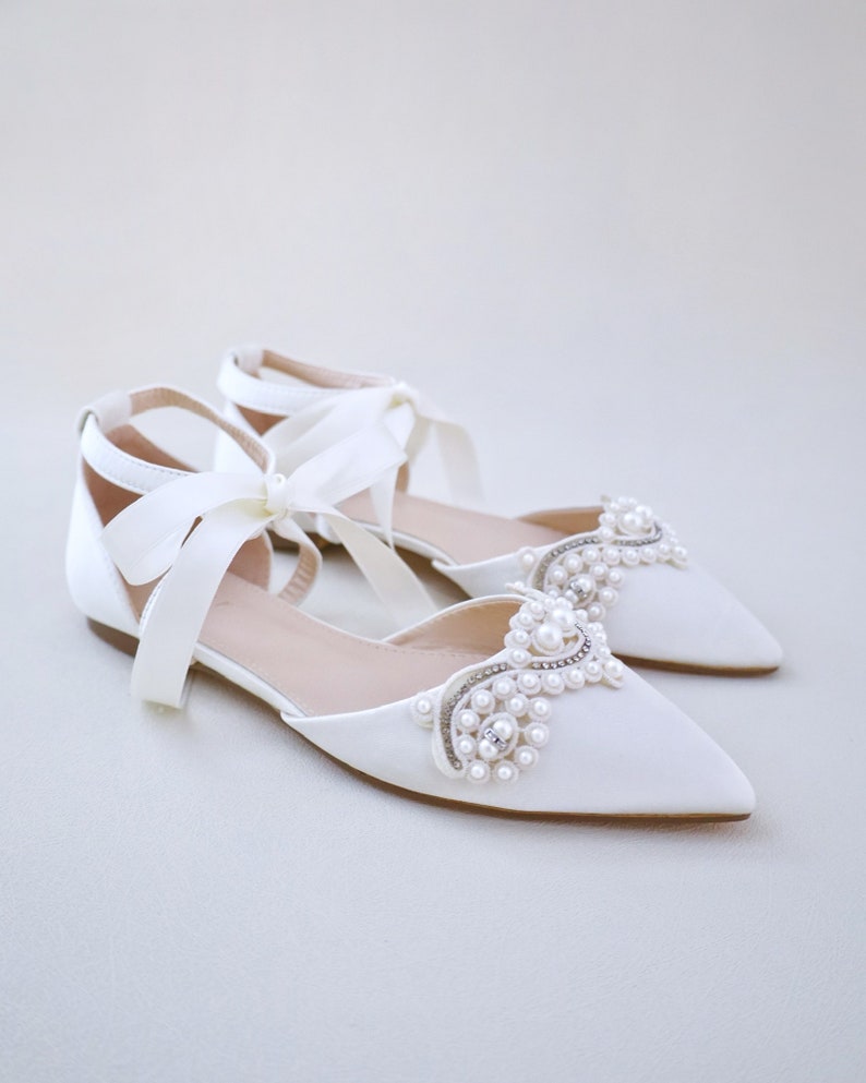 May include: A pair of ivory satin flat shoes with pointed toes. The shoes feature a decorative arrangement of pearls and rhinestones on the vamps and ankle straps tied with satin bows. The shoes are designed for special occasions.