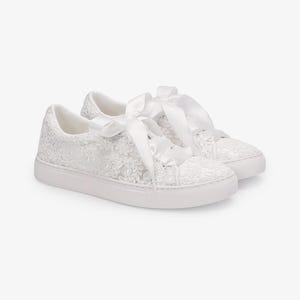 May include: A pair of white sneakers with intricate floral lace detailing. The sneakers feature white satin ribbon laces tied into bows. The soles are white and the overall design is elegant and feminine.