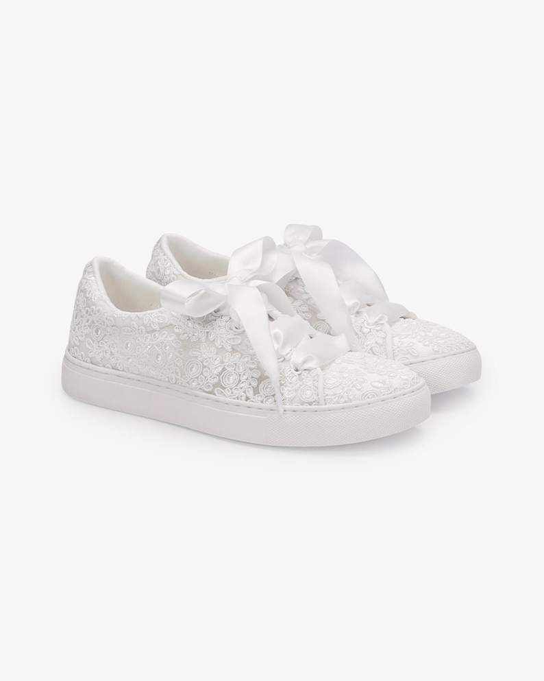 May include: A pair of white sneakers with intricate floral lace detailing. The sneakers feature white satin ribbon laces tied into bows. The soles are white and the overall design is elegant and feminine.