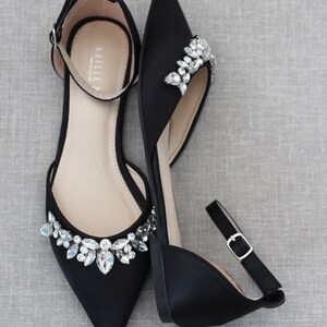 May include: A pair of black satin flats with ankle straps and crystal embellishments. The shoes have a pointed toe and a low heel.
