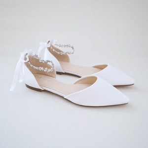 May include: A pair of white satin ballet flats with pointed toes. The ankle straps are adorned with sparkling rhinestones and tied with delicate white ribbons. The shoes are designed for special occasions.
