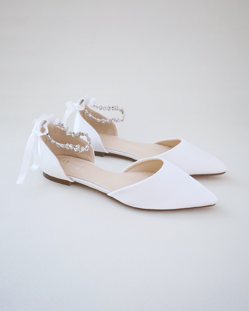 May include: A pair of white satin ballet flats with pointed toes. The ankle straps are adorned with sparkling rhinestones and tied with delicate white ribbons. The shoes are designed for special occasions.