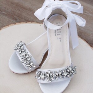 May include: White satin sandals with a rhinestone embellishment and a white satin ribbon tie. The sandals have a low heel and are perfect for a special occasion.