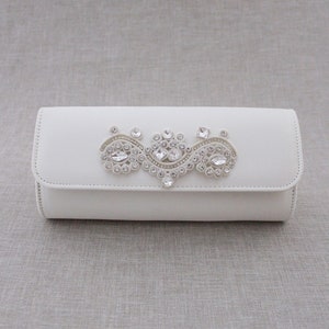 May include: A white clutch purse with a decorative crystal embellishment.