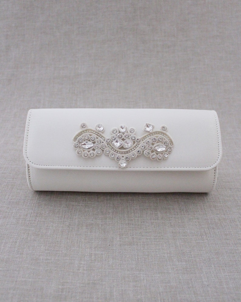 May include: A white clutch purse with a decorative crystal embellishment.