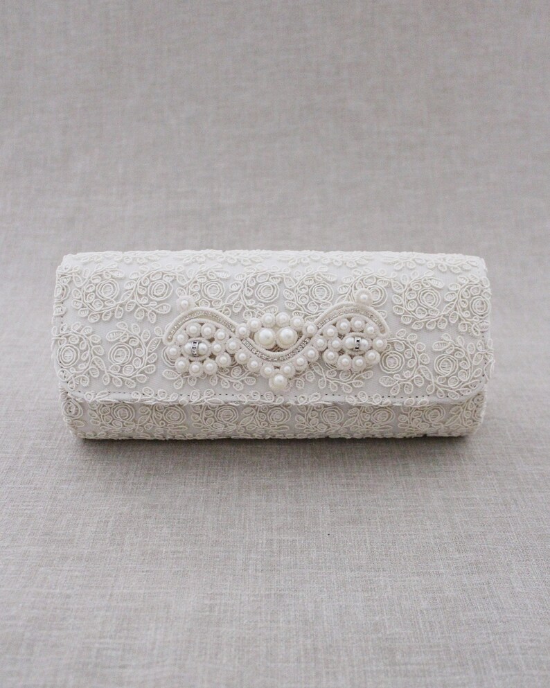 May include: A white clutch purse with a delicate floral lace design and a pearl embellishment.