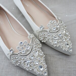 May include: Pair of white satin bridal flats with pointed toes. Each shoe features a large, ornate embellishment of clear rhinestones and crystals.