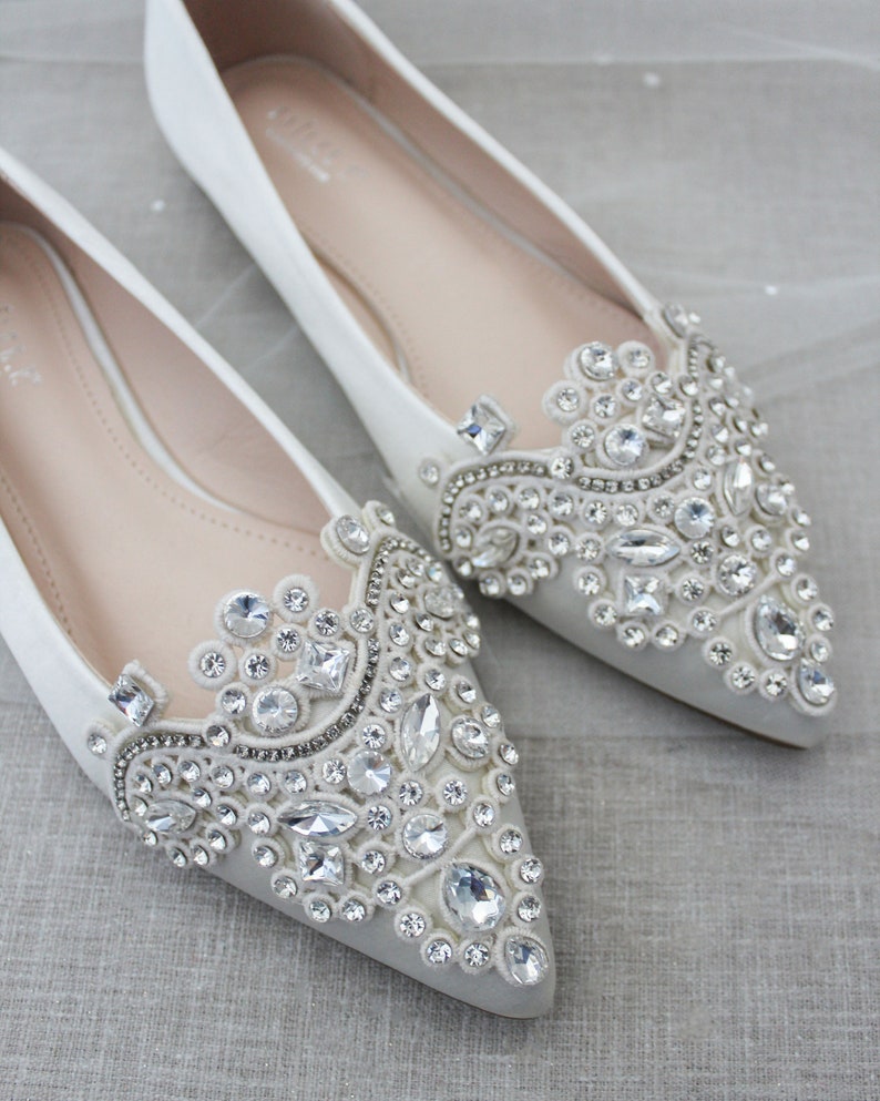 May include: Pair of white satin bridal flats with pointed toes. Each shoe features a large, ornate embellishment of clear rhinestones and crystals.