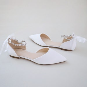May include: A pair of white satin flats with pointed toes. Each shoe features a delicate ankle strap adorned with sparkling rhinestones and a satin ribbon tie. The shoes are designed for a formal occasion.