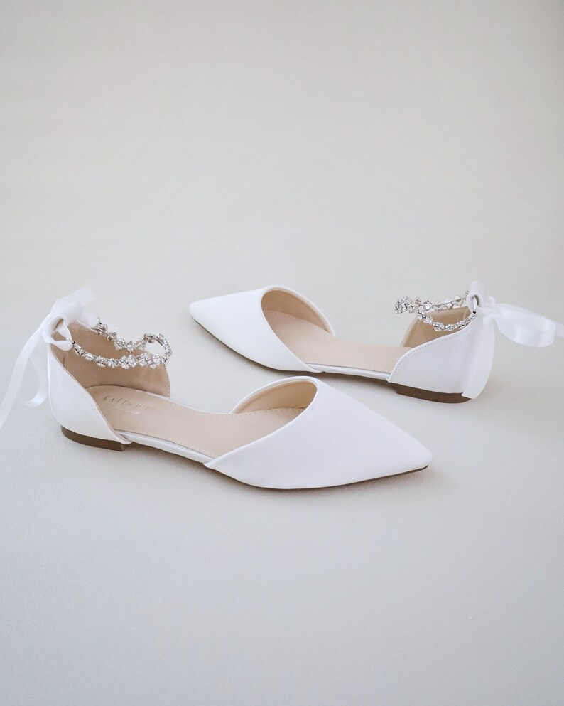 May include: A pair of white satin flats with pointed toes. Each shoe features a delicate ankle strap adorned with sparkling rhinestones and a satin ribbon tie. The shoes are designed for a formal occasion.