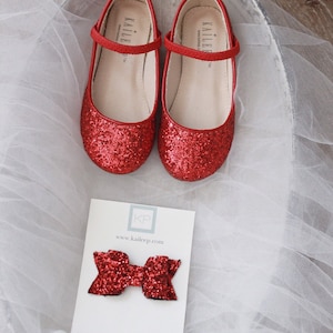 Puede incluir: Zapatillas de ballet rojas con purpurina, con una tira y la palabra "Kailee" en la tira. Los zapatos est&aacute;n sobre un fondo de tul blanco. Un lazo rojo con purpurina est&aacute; sobre una tarjeta blanca con el texto "KP www.kaileep.com".