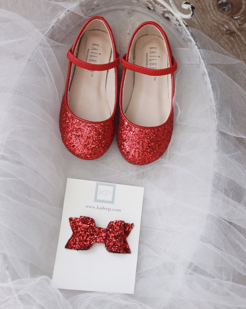 Puede incluir: Zapatillas de ballet rojas con purpurina, con una tira y la palabra "Kailee" en la tira. Los zapatos est&aacute;n sobre un fondo de tul blanco. Un lazo rojo con purpurina est&aacute; sobre una tarjeta blanca con el texto "KP www.kaileep.com".
