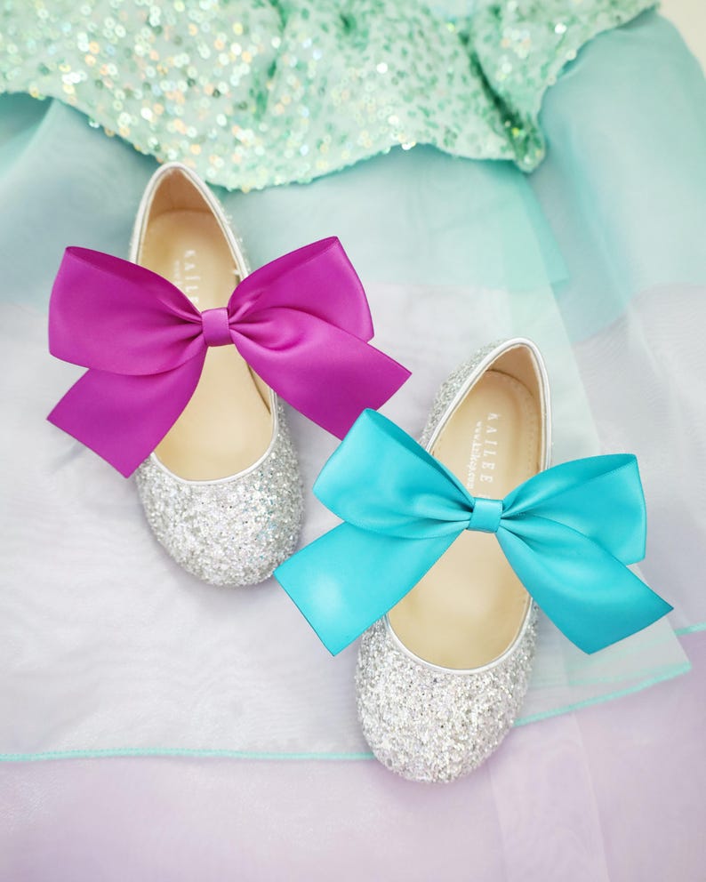 May include: Two pairs of silver glitter shoes with satin bows. One pair has a bright pink bow and the other pair has a turquoise bow.