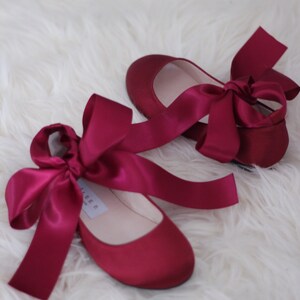 May include: A pair of burgundy satin ballet flats with matching satin ribbon ties. The ribbons are tied into bows at the front of the shoes.