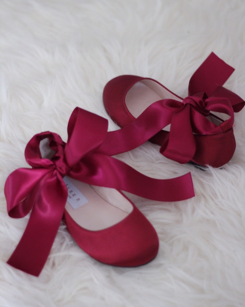 May include: A pair of burgundy satin ballet flats with matching satin ribbon ties. The ribbons are tied into bows at the front of the shoes.