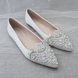 May include: A pair of ivory satin bridal flats with pointed toes. The shoes are embellished with a large, intricate design of clear rhinestones.