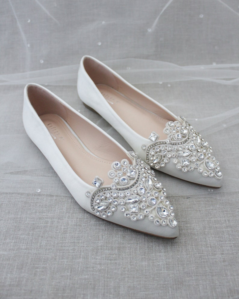 May include: A pair of ivory satin bridal flats with pointed toes. The shoes are embellished with a large, intricate design of clear rhinestones.