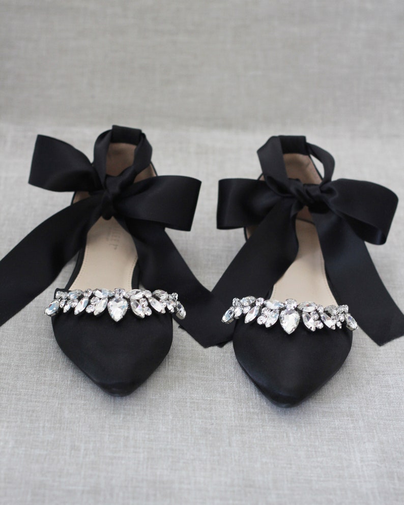 May include: Pair of black satin flats with a pointed toe and a large black satin bow on each shoe. The shoes are adorned with a row of clear rhinestones.