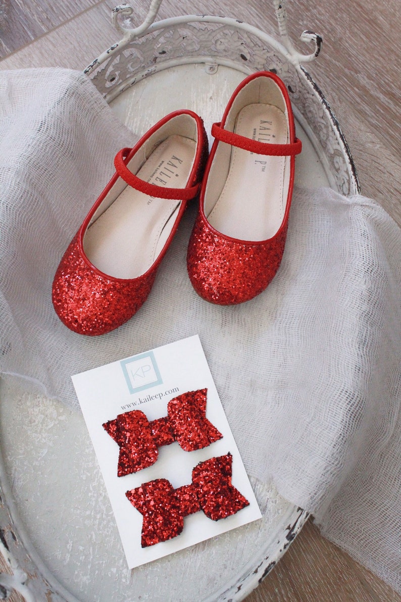 Puede incluir: Dos pares de lazos para el pelo de purpurina roja en una tarjeta blanca con el texto "KP www.kaileep.com". Los lazos est&aacute;n en una bandeja blanca con dos zapatos rojos de purpurina.