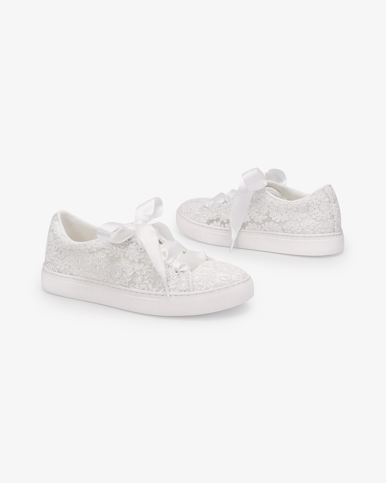 May include: A pair of white lace sneakers with white ribbon laces. The sneakers have a low profile, white rubber sole. The lace detailing covers the entire shoe, creating a delicate and elegant look. The sneakers are set against a plain white background.