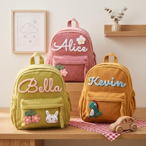 Puede incluir: Tres mochilas de pana en verde oliva, rosa empolvado y amarillo mostaza. Cada mochila tiene un asa superior, un bolsillo frontal con cremallera y un nombre personalizado en letra cursiva. Los parches decorativos incluyen flores, un conejo y un dinosaurio.