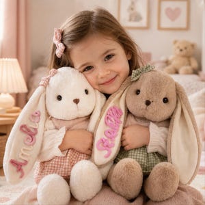 May include: Two plush stuffed rabbits with long floppy ears. The white rabbit has the name Amelia embroidered in pink on its ear, and the brown rabbit has the name Stella embroidered in pink on its ear. Both rabbits wear small checkered outfits.